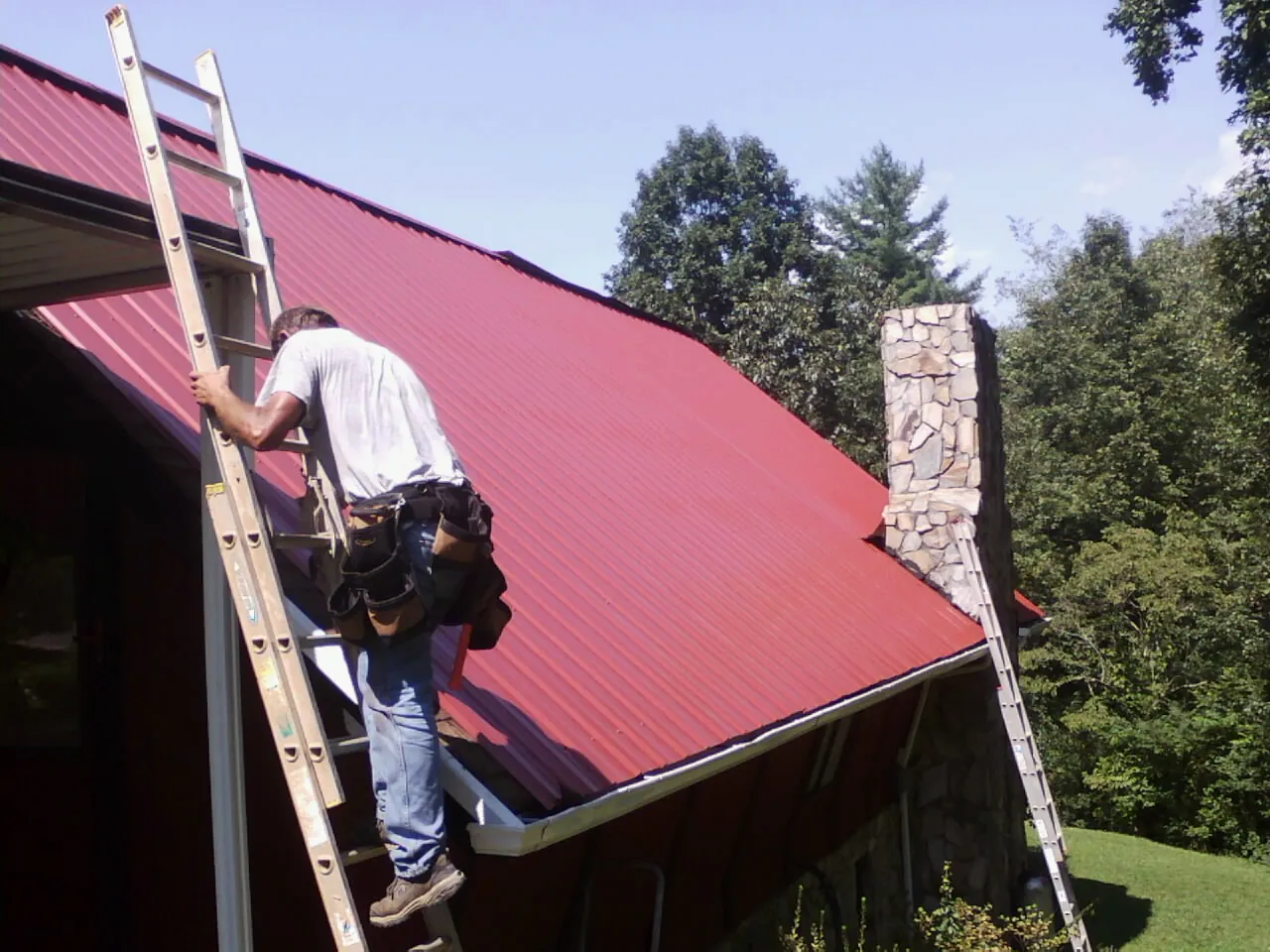 Roofer on a ladder beside a red metal roof and stone chimney — Soffit & Fascia Repair by Roofing Brothers in Potts Camp